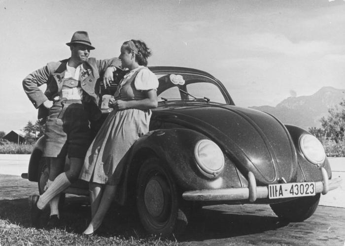 A man and a woman in traditional German dress lean against a dark colored Volkswagen car. A mountain range appears in the background.